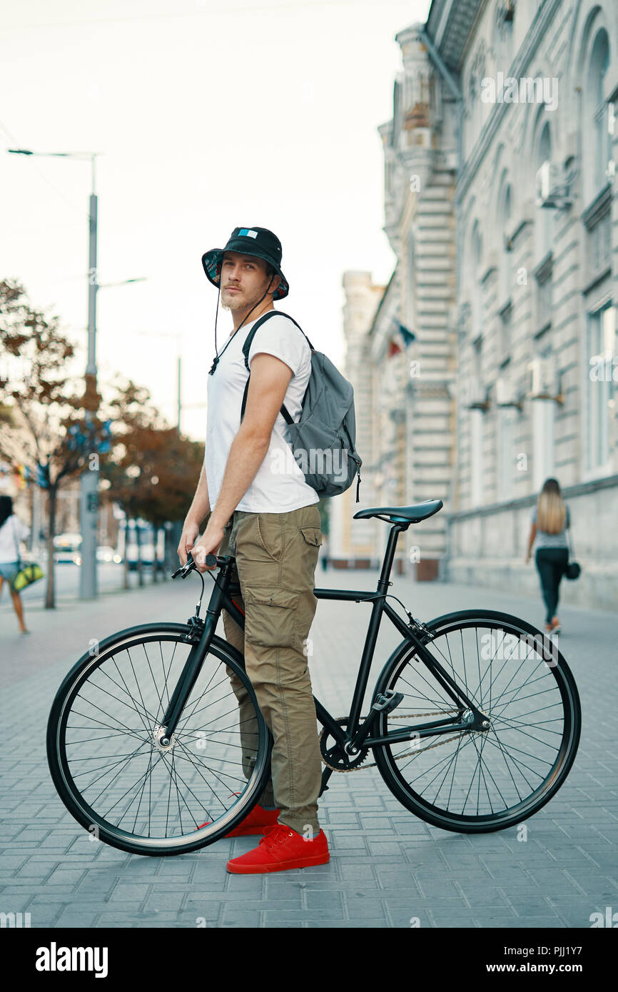 Outdoor ritratto di un moderno e giovane uomo della strada, seduto sulla  bici. Un giovane uomo di atletica che indossa i pantaloni khaki, T-shirt  bianco, rosso sneakers e backp Foto stock -