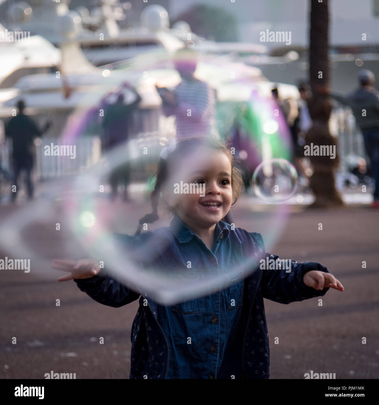 Bambina è felice di gigantesche bolle di sapone, sorriso luminoso, persone e marina in background Foto Stock