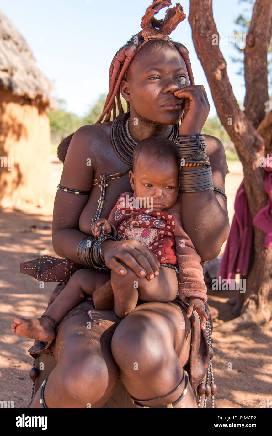 Himba donna siede nell'ombra di un albero con il bambino in braccio Foto Stock