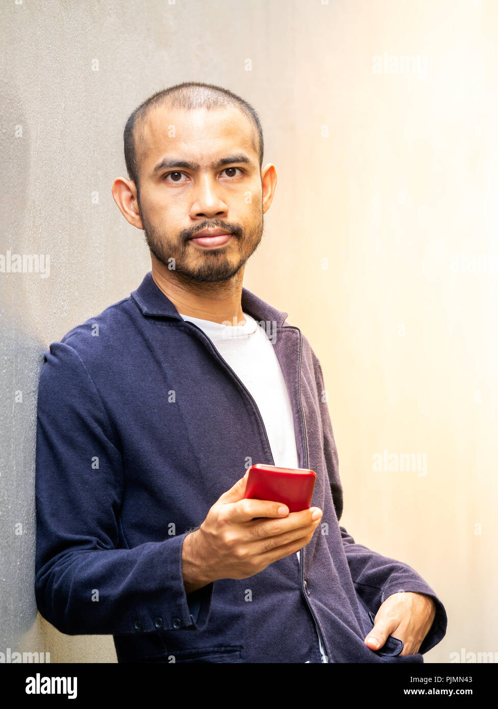Smart uomo asiatico indossando maglietta blu appoggiarsi contro la parete dello smartphone holding guardando avanti con fiducia. Foto Stock