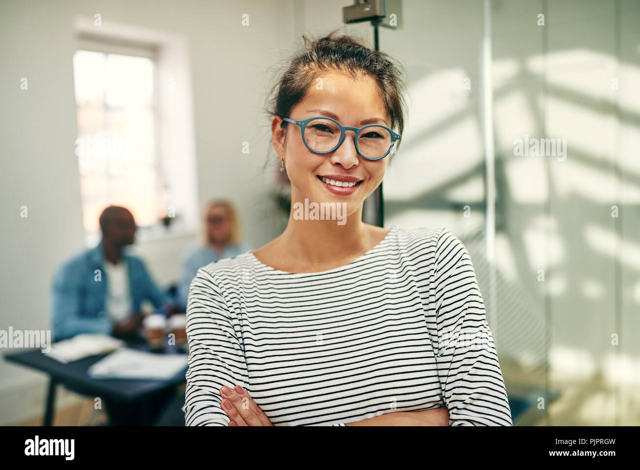 Sorridente giovane imprenditrice asiatica con gli occhiali in piedi in un ufficio con le braccia incrociate con i colleghi che lavorano in background Foto Stock
