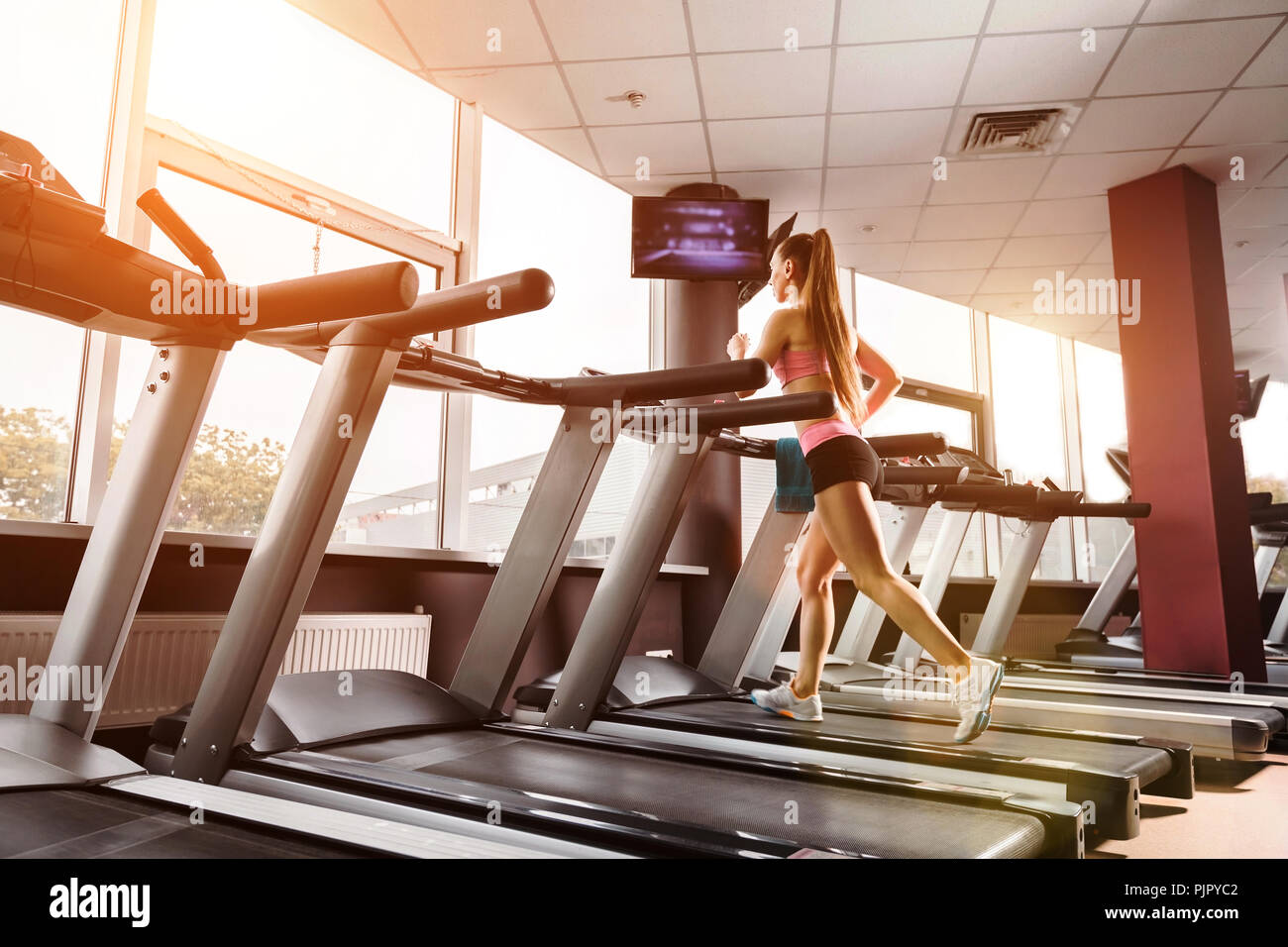 Slim Lady piuttosto acceso al tapis roulant in palestra contro il tramonto. Concetto di esercizi cardio e sano stile di vita Foto Stock