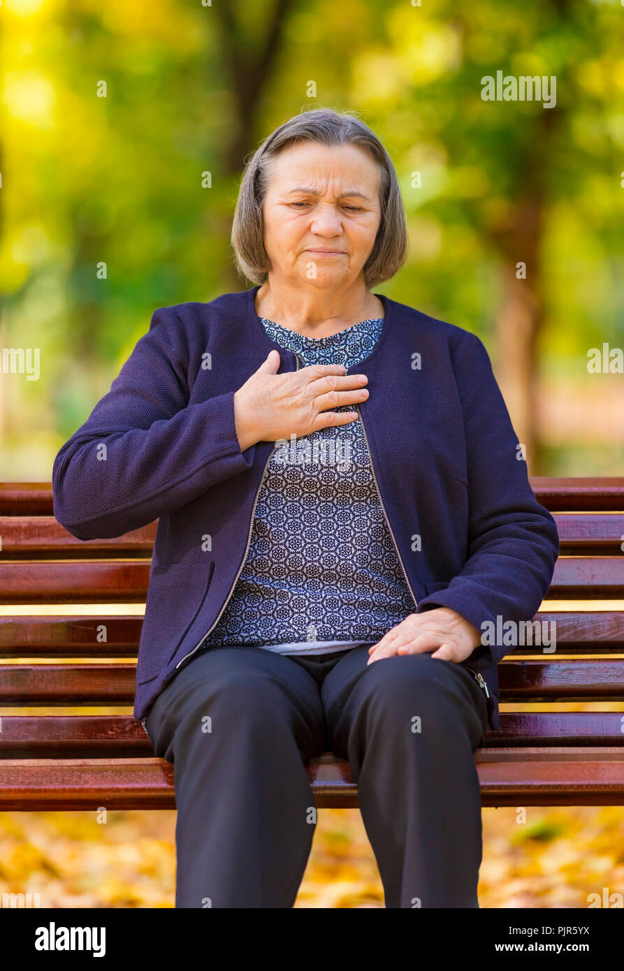 Senior lady stringendo il suo torace nel dolore ai primi segni di angina o un infarto del miocardio o un attacco di cuore, corpo superiore nel parco su una giornata autunnale Foto Stock