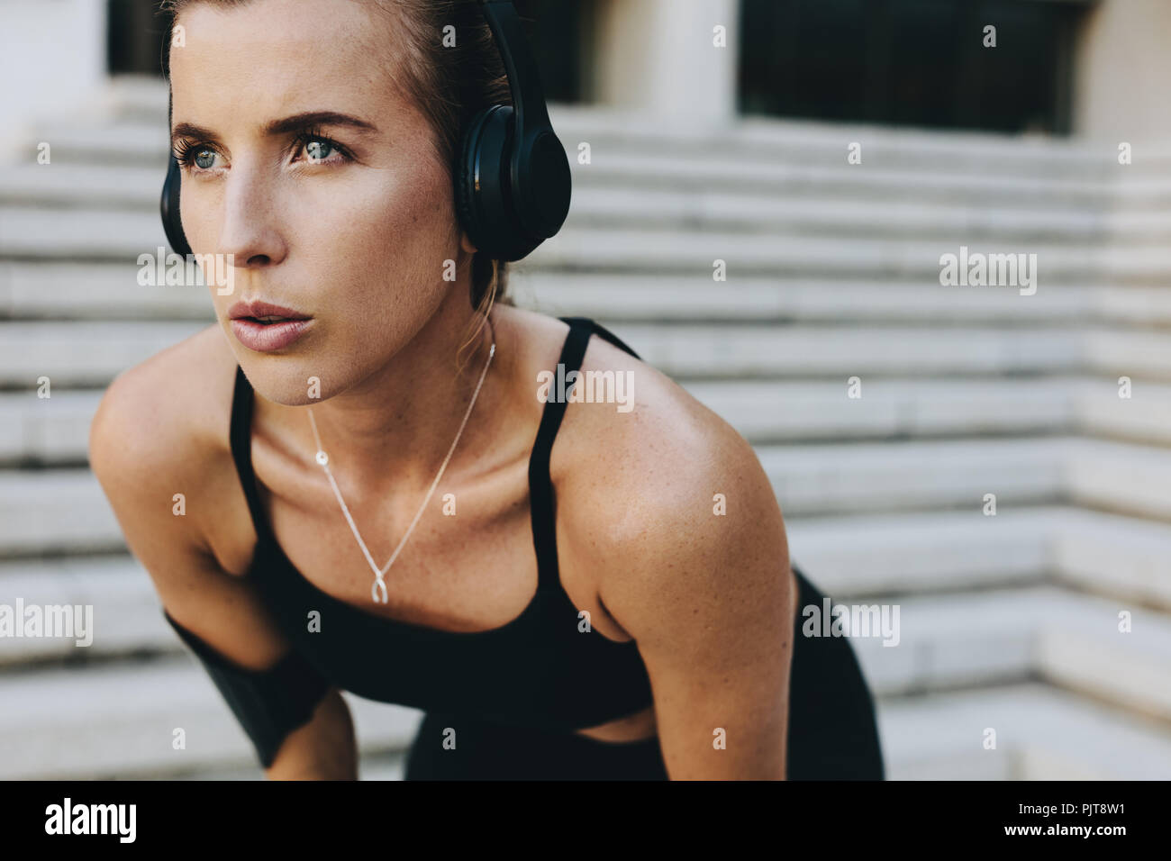 Primo piano di una donna atleta in abiti da allenamento in piedi all'aperto con le mani sulle ginocchia. donna in forma fisica che indossa le cuffie wireless per rilassarsi mentre traini Foto Stock