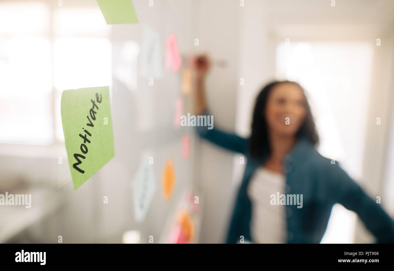 Immagine di sfocatura di un imprenditrice incollando sticky notes sulla parete di vetro con la messa a fuoco su un post-it motivare scritto su di esso. Foto Stock