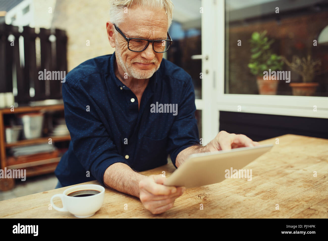 Sorridente uomo senior bevendo una tazza di caffè e di lavorare con una tavoletta digitale durante i momenti di relax in un tavolo esterno sul suo patio Foto Stock