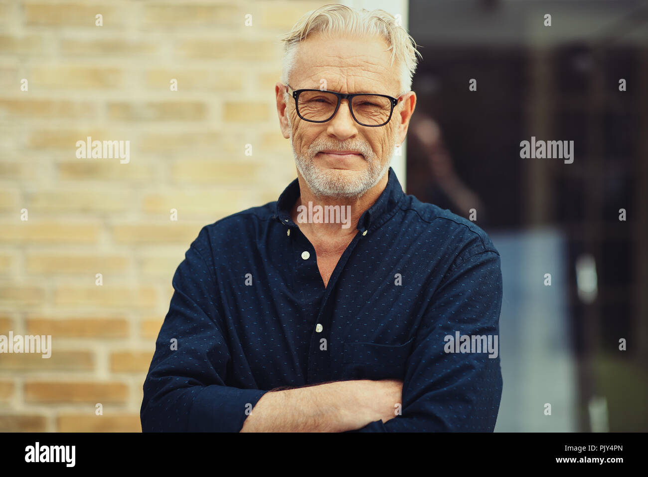 Sorridente senior con barba e con gli occhiali in piedi con le braccia incrociate al di fuori davanti a casa sua Foto Stock