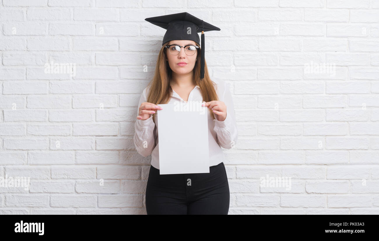 Giovane donna adulta su bianco muro di mattoni che indossa il cappuccio laureati grado di contenimento con un fiducioso espressione sul viso intelligente pensare grave Foto Stock