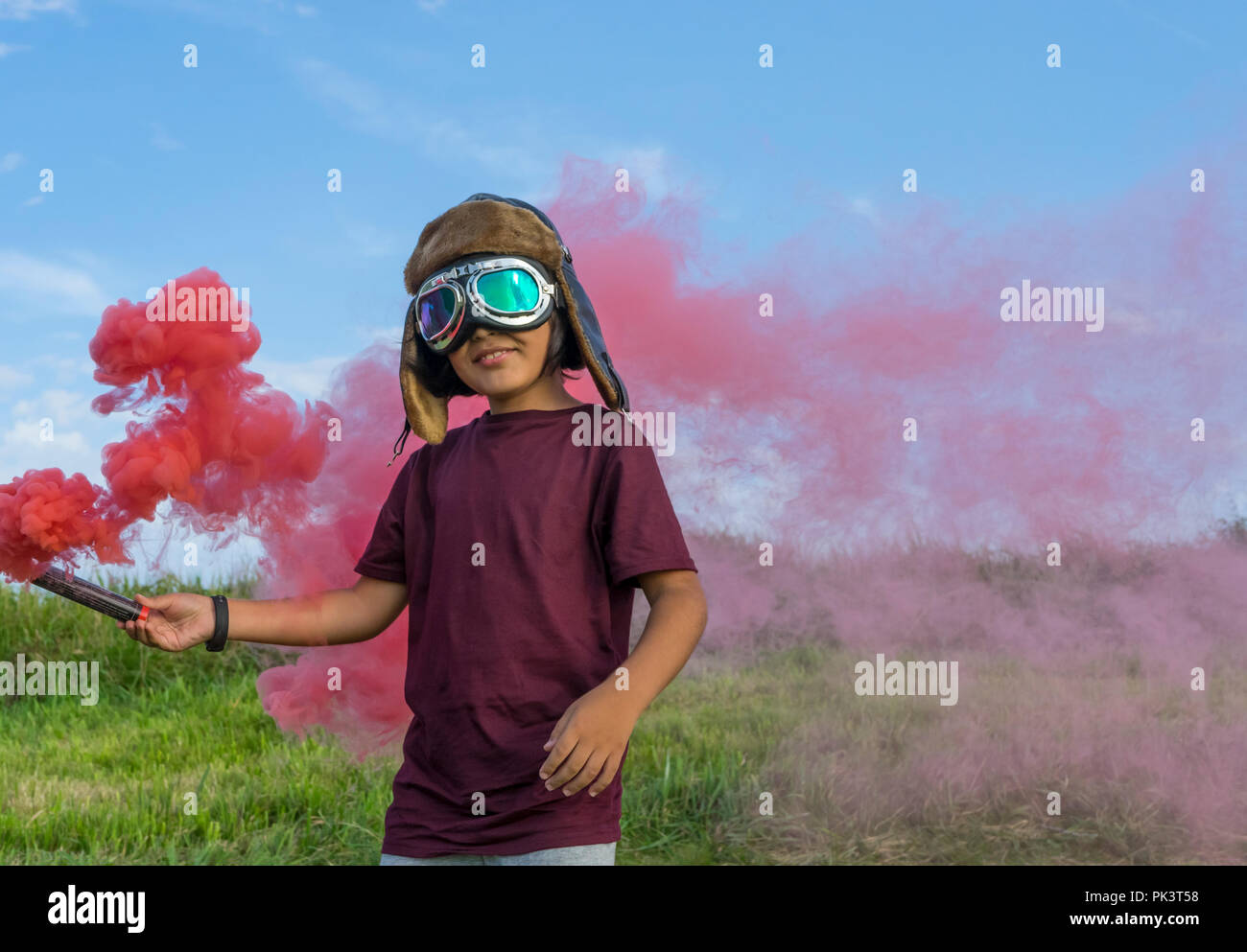 Piccolo ragazzo che indossa il casco e aeromobili googles in piedi su un campo verde con fumo colorato, fingendo di essere un pilota Foto Stock