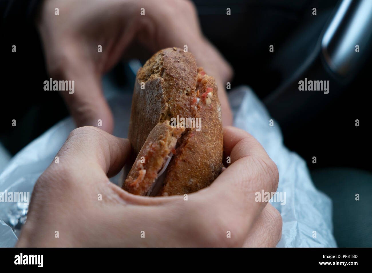 Primo piano di un giovane uomo caucasico mangiare un prosciutto e formaggio sandwich, da un sacchetto di plastica, seduti al posto di guida di una vettura Foto Stock