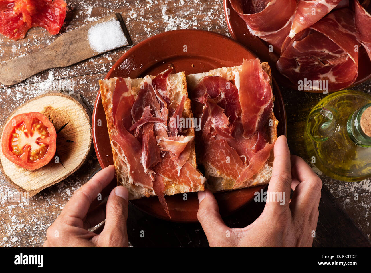 Elevato angolo di visione di un uomo la preparazione di un tipico spagnolo bocadillo de jamon, un prosciutto Serrano sandwich, su una tavola in legno rustico, accanto ad una piastra con alcuni s Foto Stock