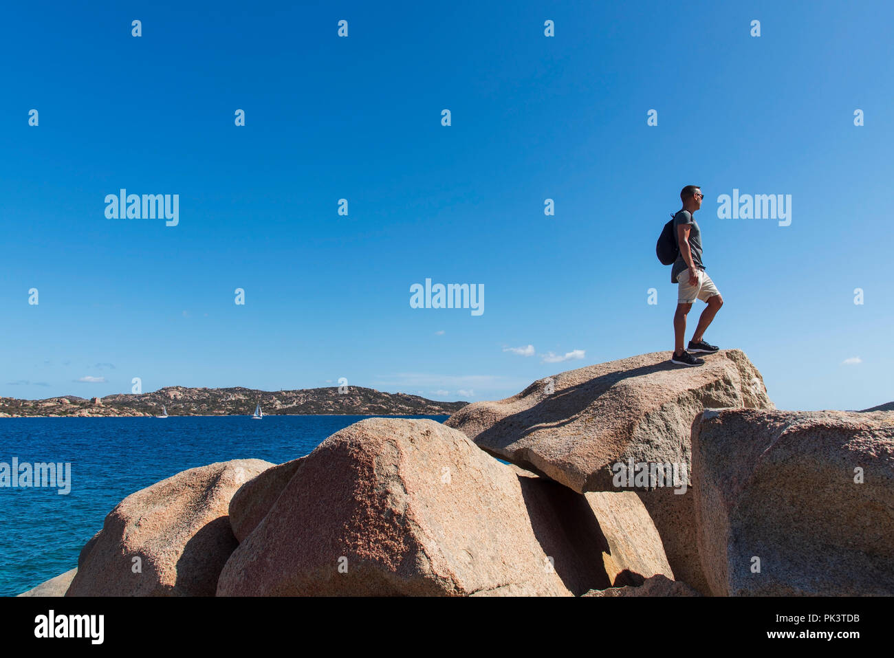 Primo piano di un giovane uomo caucasico portando uno zaino, nella parte superiore di una formazione di roccia, guardando il mare presso la Costa Smeralda, in Sardegna, Italia Foto Stock