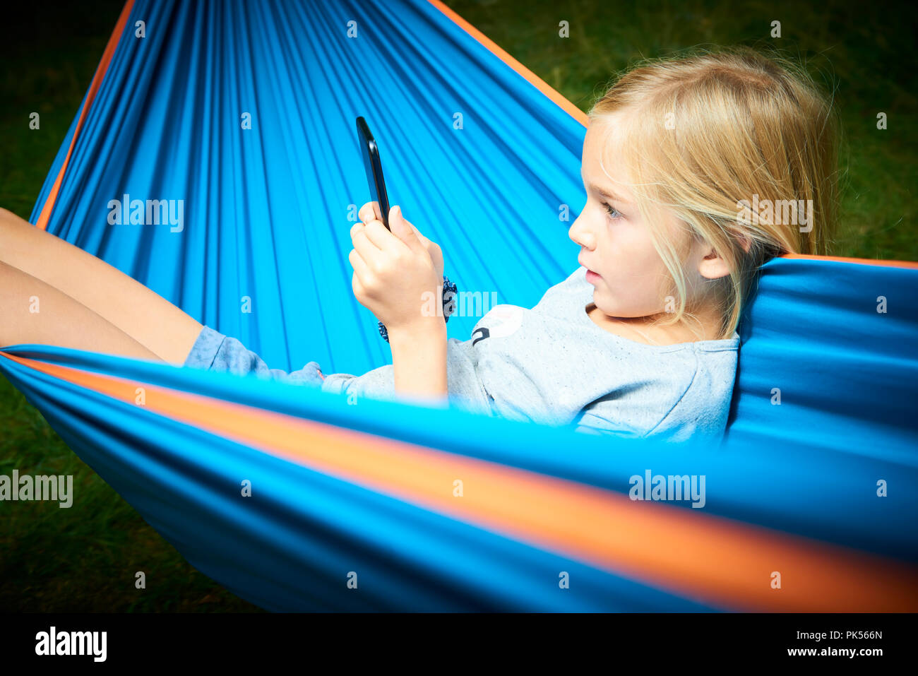 Bambino ragazza bionda giocando con uno smart phone sdraiati sulle amache all'aperto Foto Stock