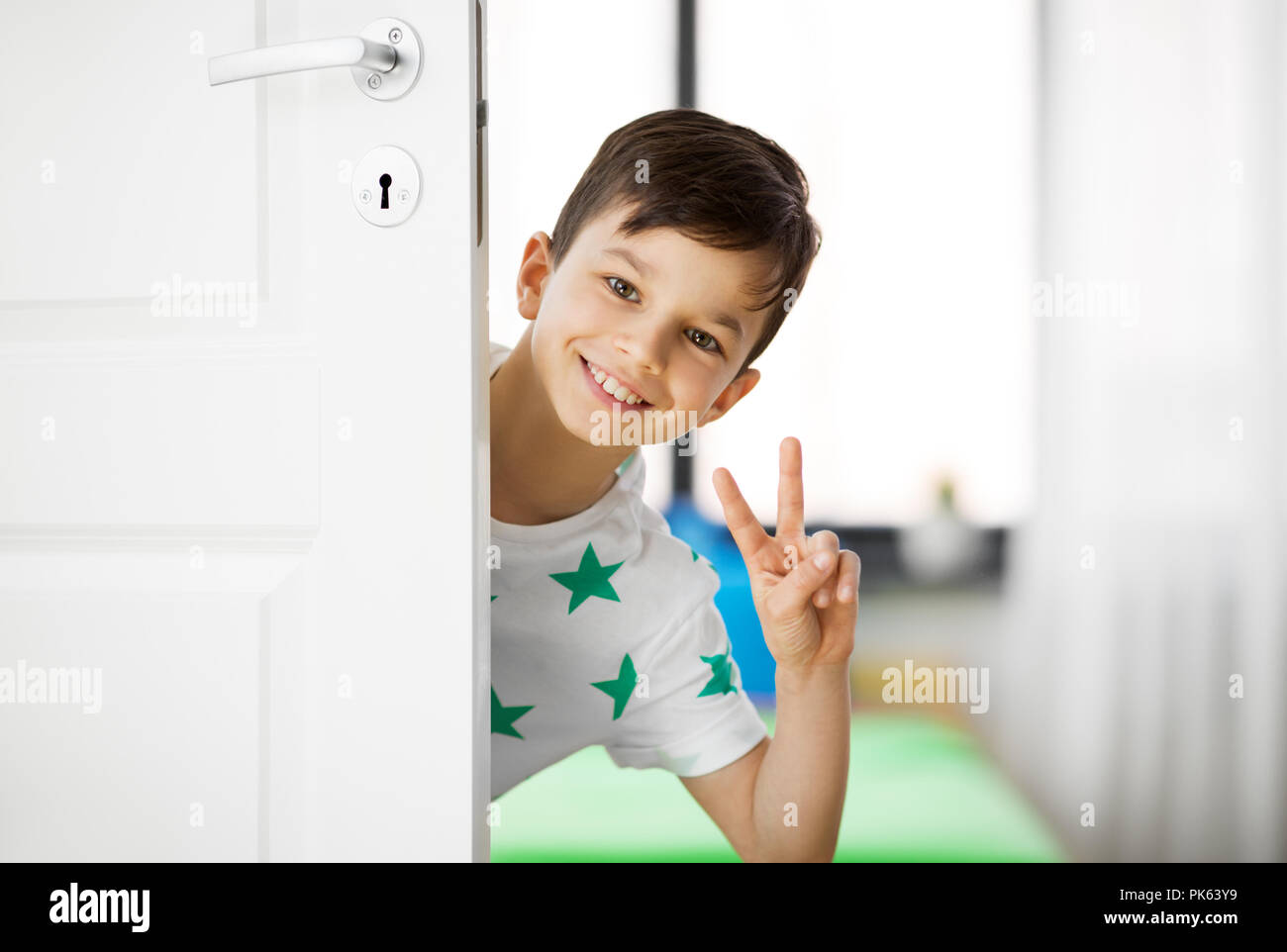Happy little boy dietro la porta di casa che mostra la pace Foto Stock
