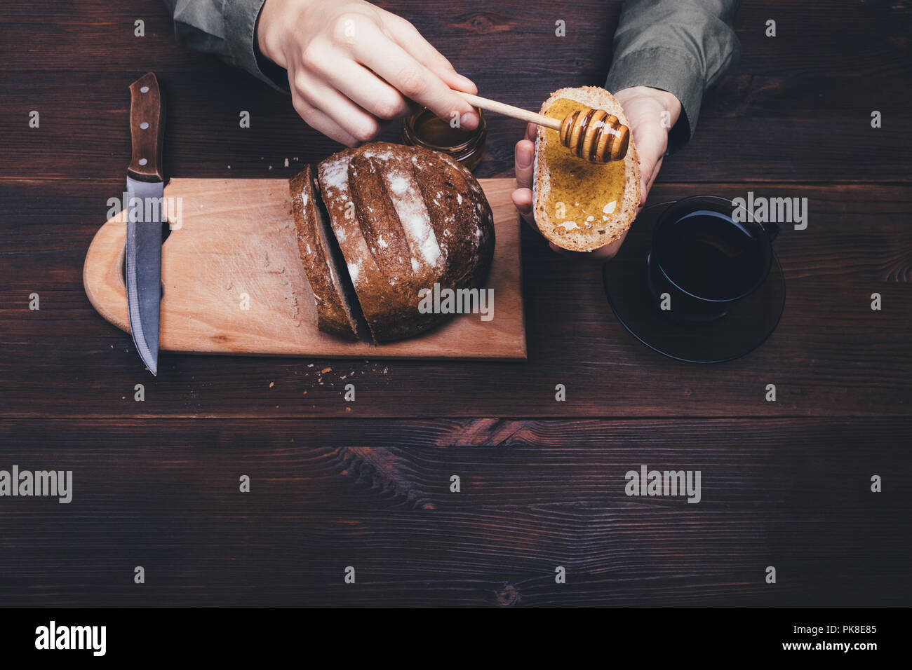 Vista superiore mani femminili fanno un sandwich di pane di segale e miele scuro sul tavolo di legno accanto alla tazza di tè nero o caffè. Composizione di semplici piatti rustici. Foto Stock