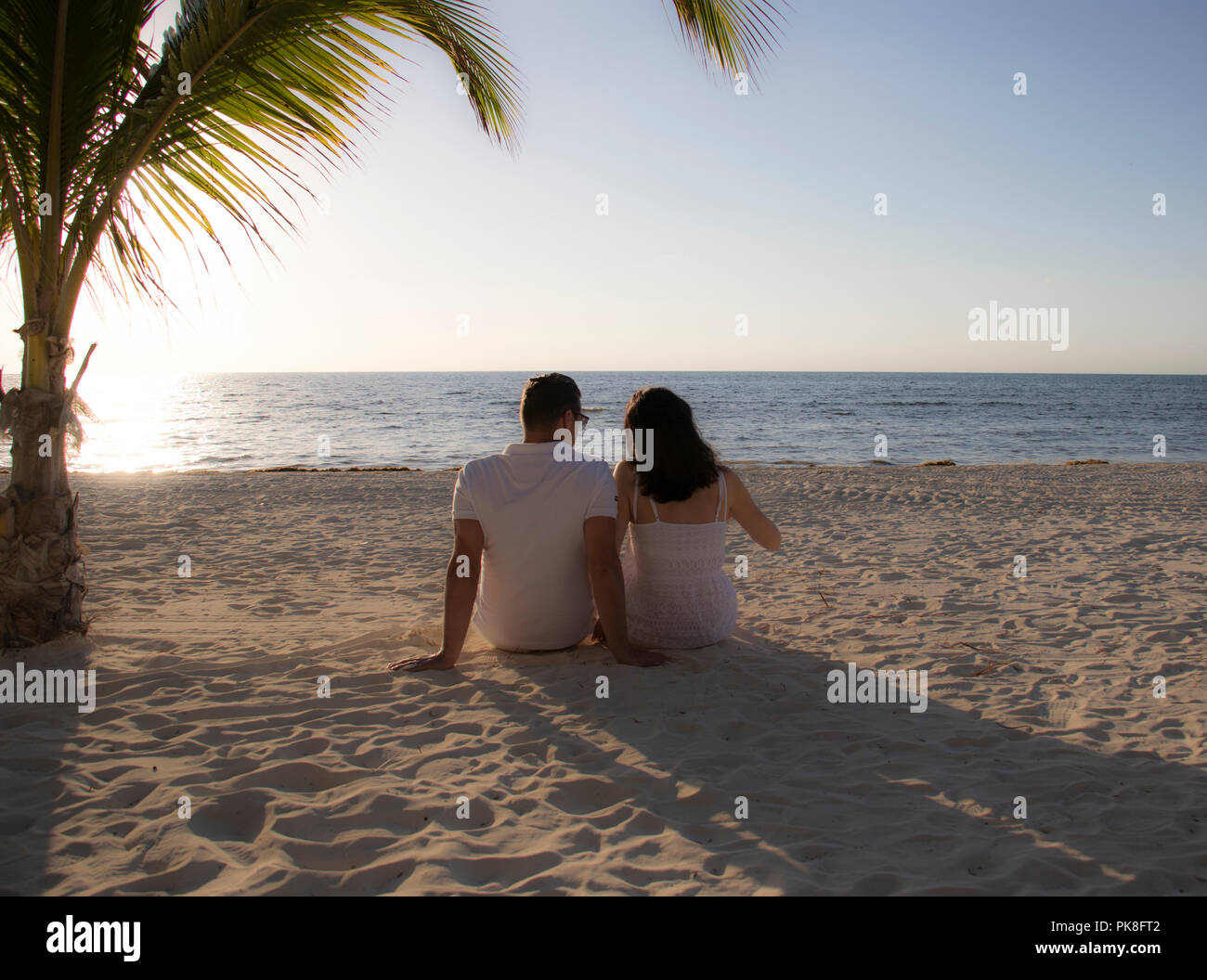 Carino coppia guardando il tramonto su una spiaggia con palme. Retro del giovane seduta nella sabbia su una spiaggia durante una vacanza tropicale. Foto Stock