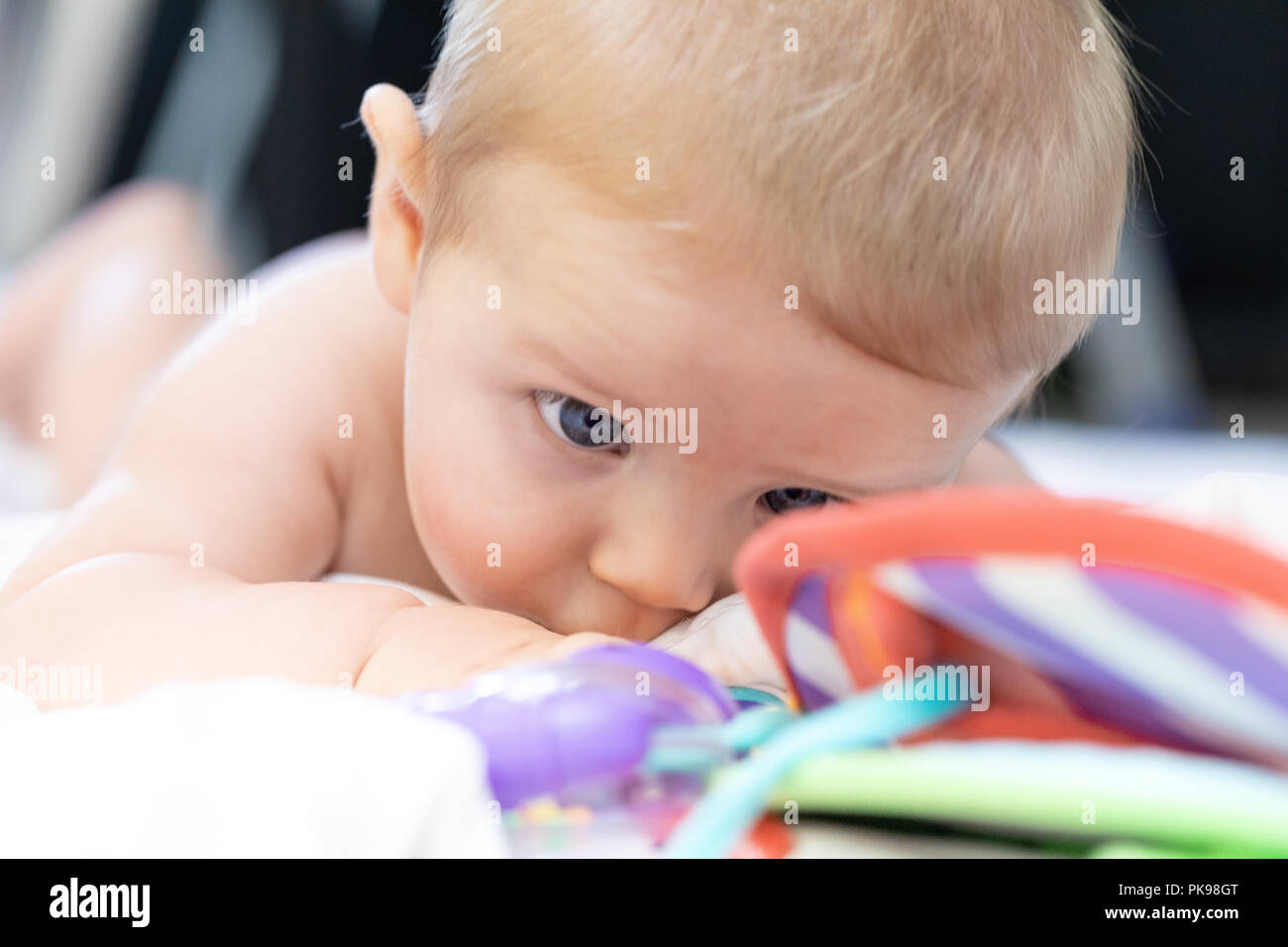 Piccolo neonato occhiatura i suoi colorati giocattoli ammassati nelle cot come esso giace sul suo stomaco su lenzuola bianche in una stretta fino a basso angolo di visione Foto Stock