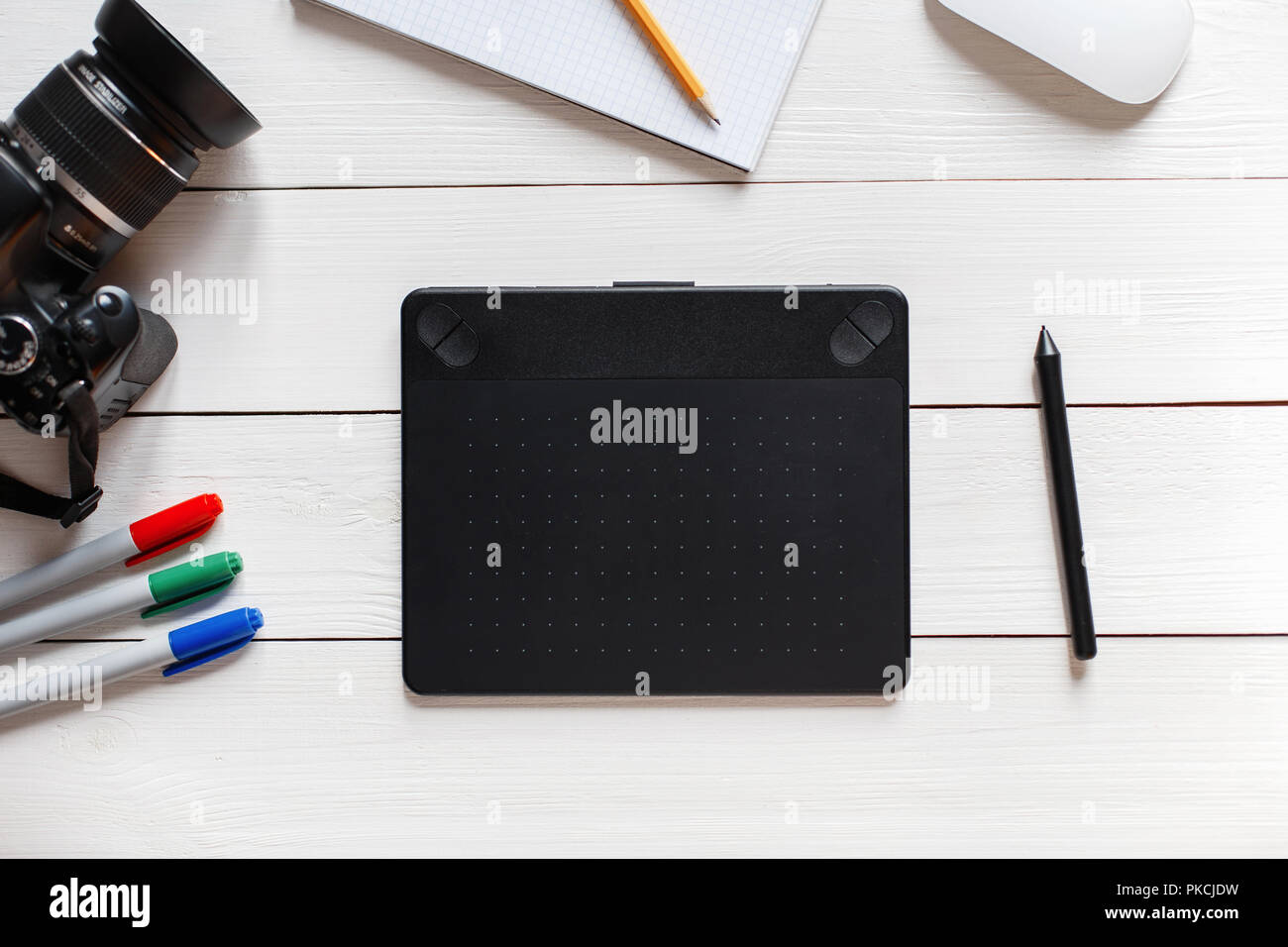 Concetto di artista Desktop. Tavoletta grafica, fotocamera reflex, mouse, taccuino, matita e pennarelli su un bianco tavolo di legno. Vista da sopra Foto Stock