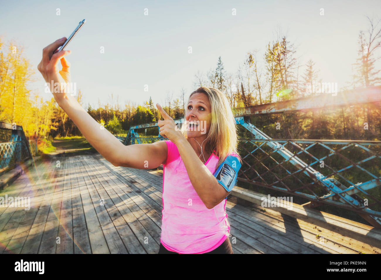 Una donna che indossa active wear e una fascia da braccio per il suo telefono cellulare si erge su un ponte in un parco in autunno prendendo un autoritratto con il suo telefono cellulare Foto Stock