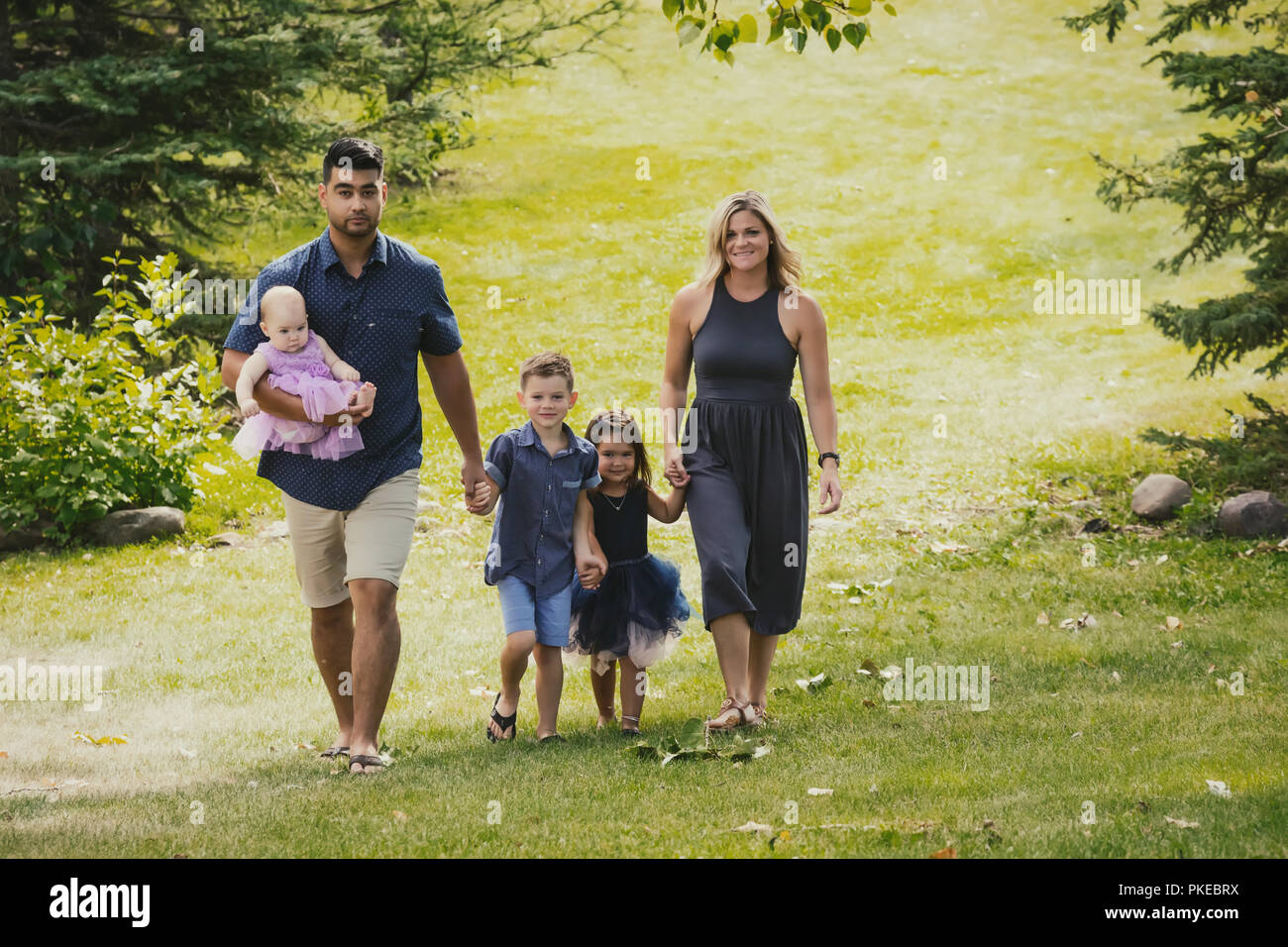 Una madre e un padre in una gara mista matrimonio a piedi con i loro figli in un parco in una gita in famiglia durante una calda giornata di autunno e il loro figlio si pone per... Foto Stock