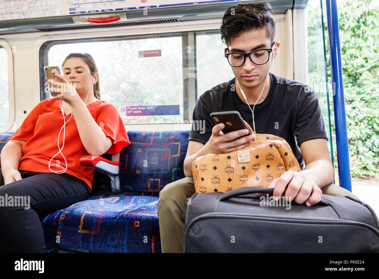Londra Inghilterra,UK,Picadilly Circus Line,treno interno uomo uomo donna donna donna maschile, seduta con smartphone auricolari passeggeri bagagli passeggeri Foto Stock