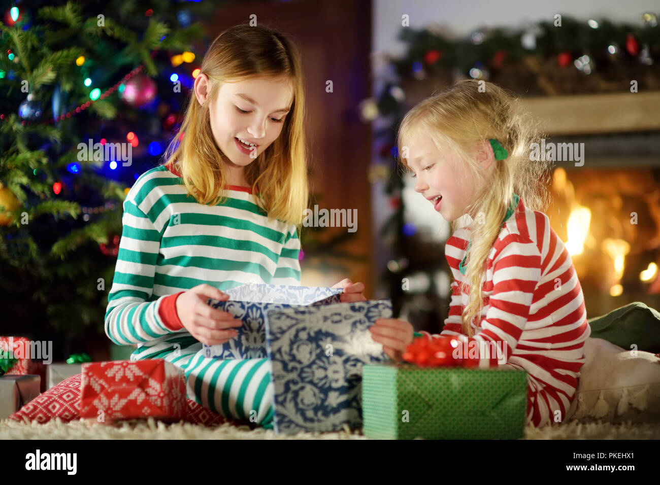 Felice piccole sorelle indossando pigiami di Natale apertura scatole regalo da un camino in un buio accogliente soggiorno alla vigilia di Natale. Celebrare il Natale a casa Foto Stock