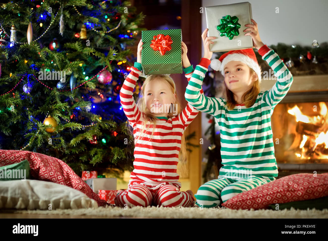 Felice piccole sorelle indossando pigiami di Natale apertura scatole regalo da un camino in un buio accogliente soggiorno alla vigilia di Natale. Celebrare il Natale a casa Foto Stock