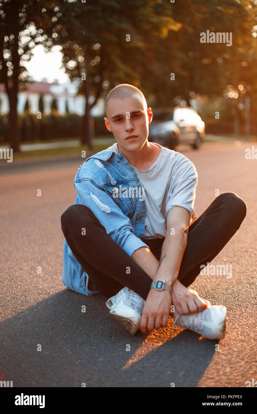 Elegante giovane uomo brutale con abbigliamento in jeans in grigio di una T- shirt, pantaloni neri, scarpe bianche si siede su una strada asfaltata al  tramonto Foto stock - Alamy