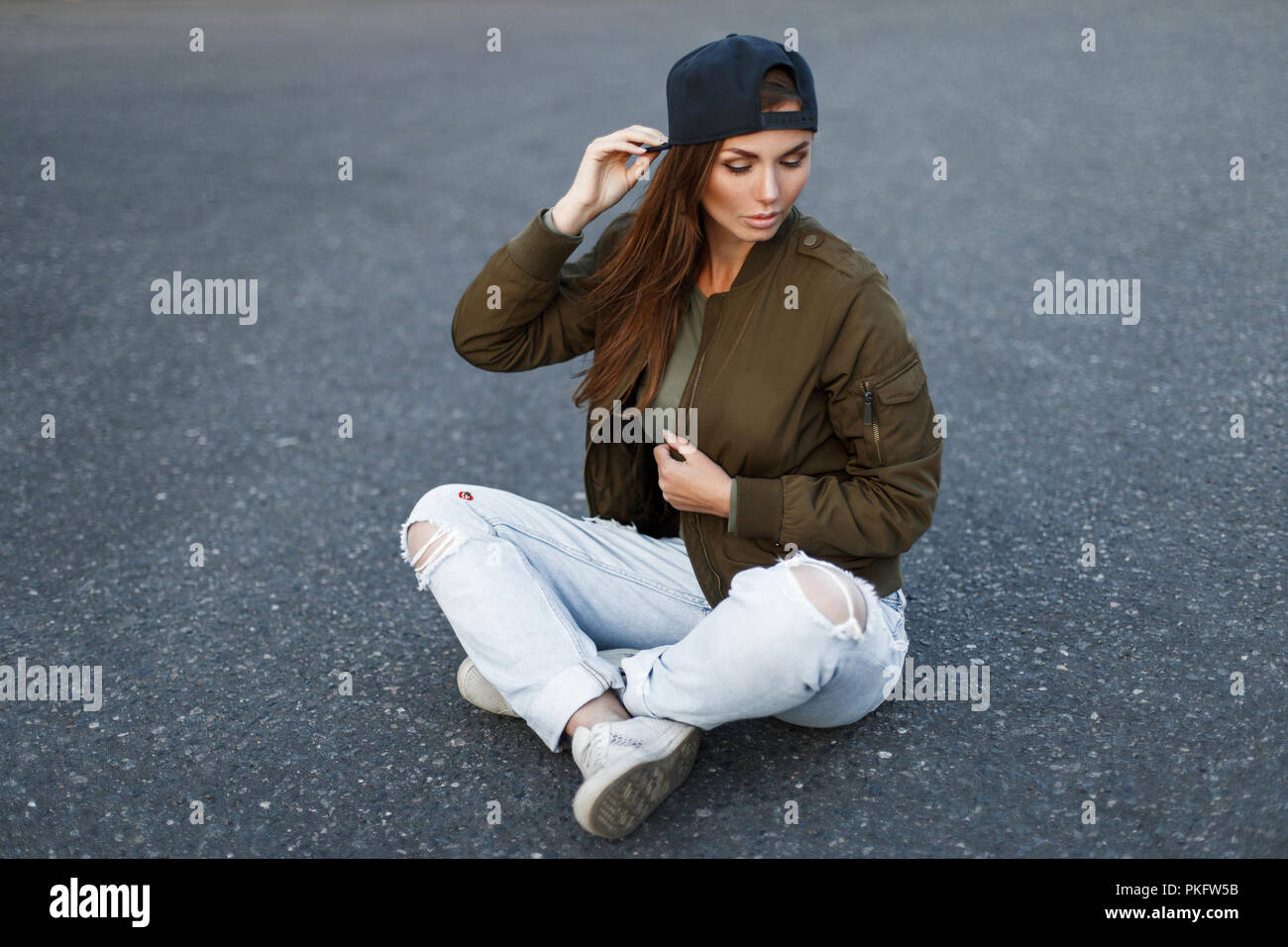 Bella giovane donna con lentiggini in un nero cappello da baseball in una  zona alla moda verde militare giacca e jeans strappati siede su asfalto  Foto stock - Alamy