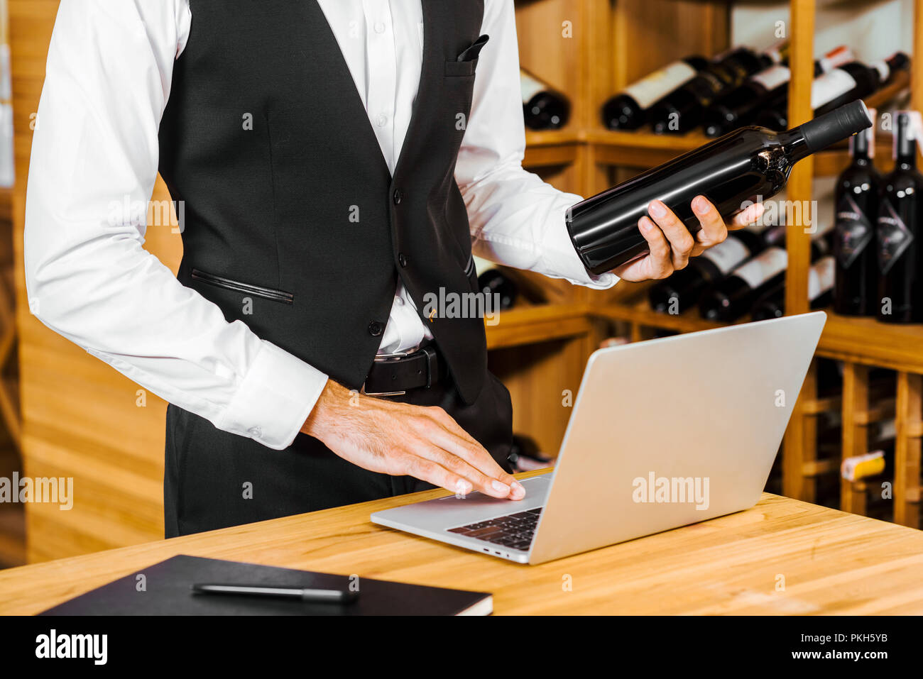 Ritagliato colpo di sommelier tenendo la bottiglia e lavorare con computer portatile presso il negozio dei vini Foto Stock