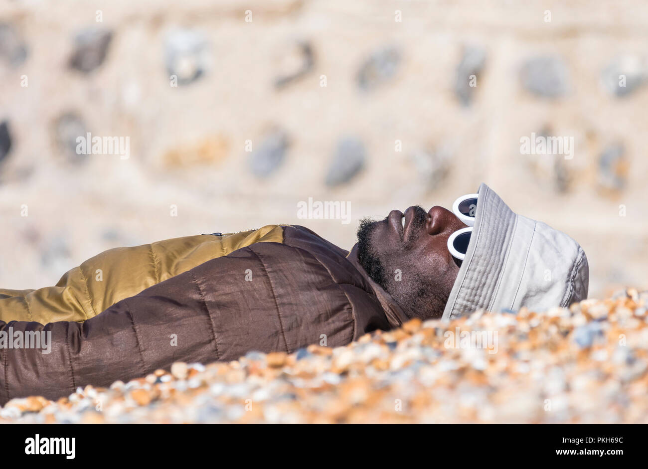 Uomo nero posa su una spiaggia in un giorno caldo e soleggiato in primavera indossa un cappello e il cappotto di occhiali da sole, nel Regno Unito. Foto Stock