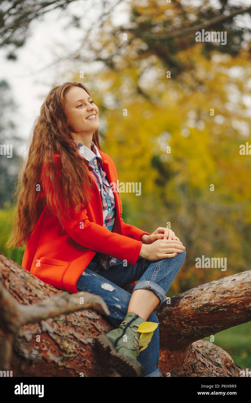 Giovane bella donna con lunghi capelli ondulati in rosso e il cappotto di jeans seduto su un ramo di albero. Ragazza sorridente ritratto d'autunno. Foto Stock