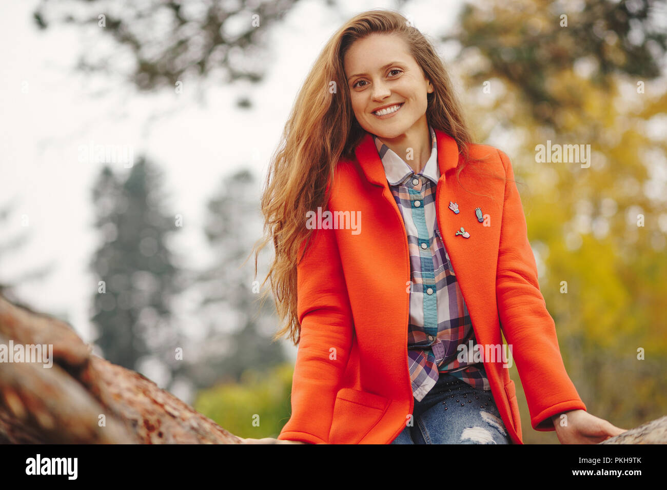 Giovane bella donna con lunghi capelli ondulati in rosso e il cappotto di jeans seduto su un ramo di albero. Ragazza sorridente ritratto d'autunno. Foto Stock
