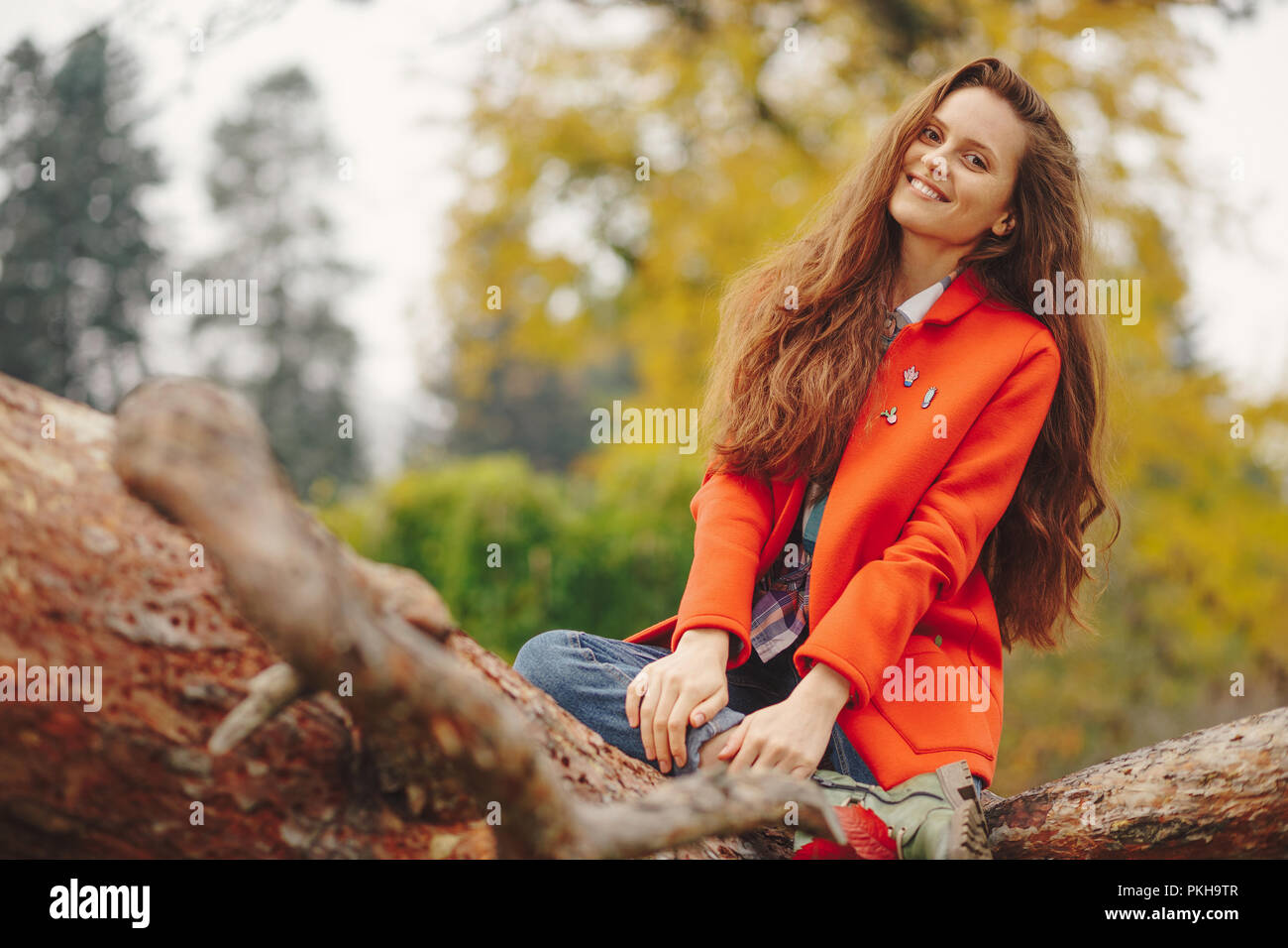 Felice bella donna con lunghi capelli ondulati in rosso e il cappotto di jeans seduto su un ramo di albero. Ragazza sorridente ritratto d'autunno. Foto Stock