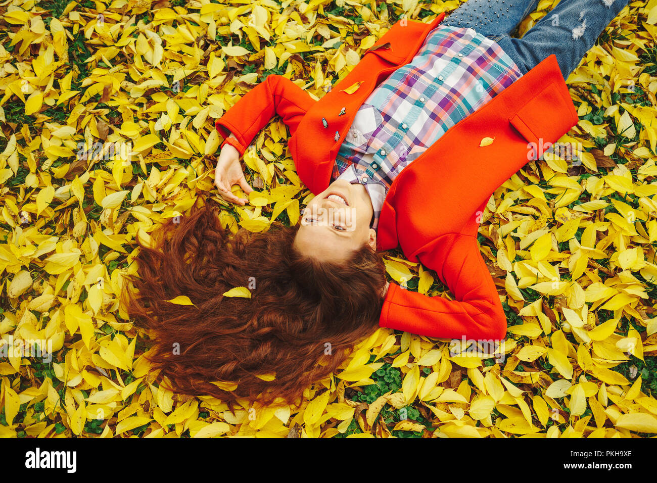 Sorridente ragazza con lunghi capelli ondulati gustano la vita nella foresta di autunno. Allegro bella ragazza in rosso cappotto e colorato plaid shirt su bella giornata di caduta. Vi Foto Stock