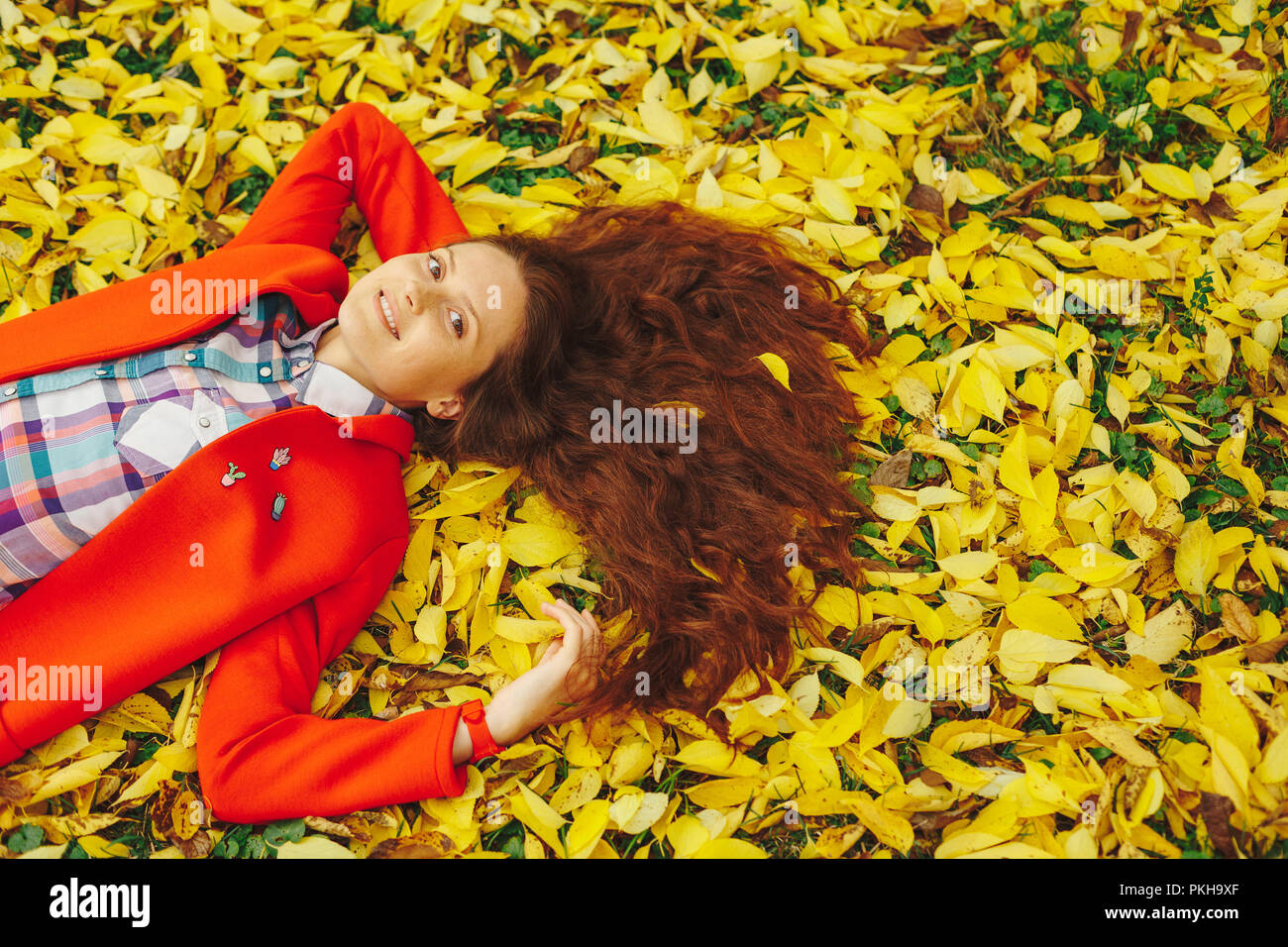 Sorridente ragazza con lunghi capelli ondulati per godersi la vita nella foresta di autunno. Allegro bella ragazza in rosso cappotto e colorato plaid shirt su bella giornata di caduta. H Foto Stock