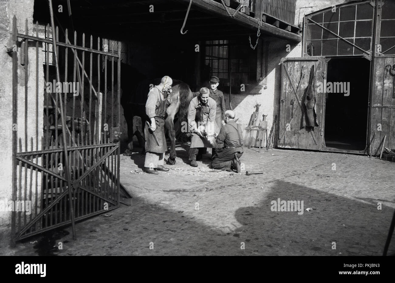 Degli anni Cinquanta, storico, maniscalchi fuori in una città interna cortile in ciottoli o stalle, lavorando su uno zoccolo di cavallo, Colonia, Germania. Un maniscalco è altamente esperto artigiano, combinando alcune abilità di fabbro con alcuni di quelli veterinario, per curare i cavalli' piedi. Foto Stock