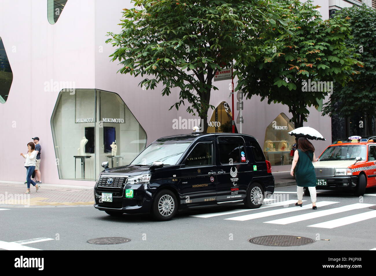 Una Toyota JPN Taxi di Tokyo Olimpiadi logo 2020 su di esso andando oltre un crosswalk nella parte anteriore della Toyo Ito-progettato Mikimoto Ginza 2 store in Tokyo. Foto Stock