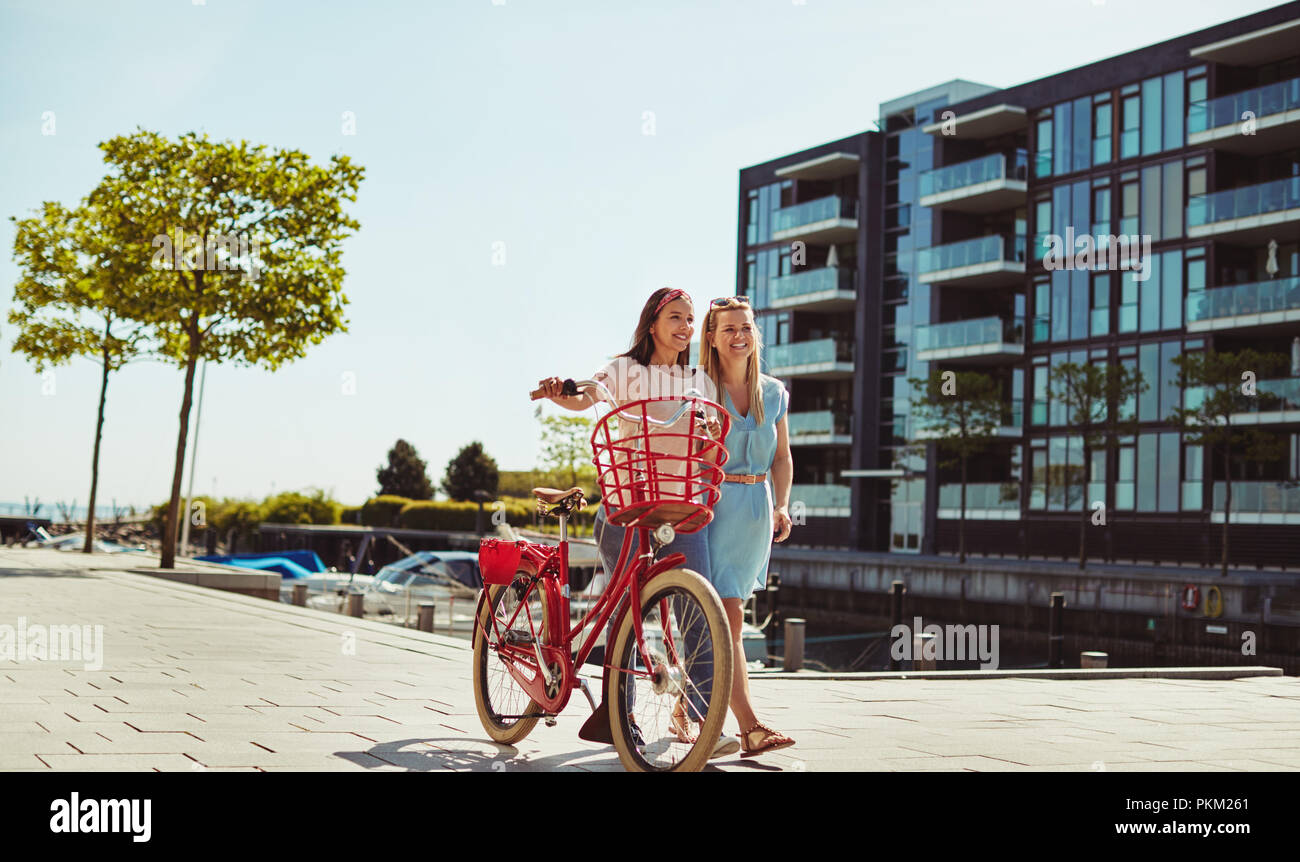 Due amiche a ridere e divertirsi mentre si cammina insieme con una bicicletta attraverso la città in estate Foto Stock
