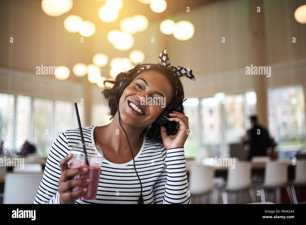 Ridendo giovane africano studente di college prendendo una pausa dallo studio mentre si beve un smoothie e ascolto di musica sulle cuffie Foto Stock