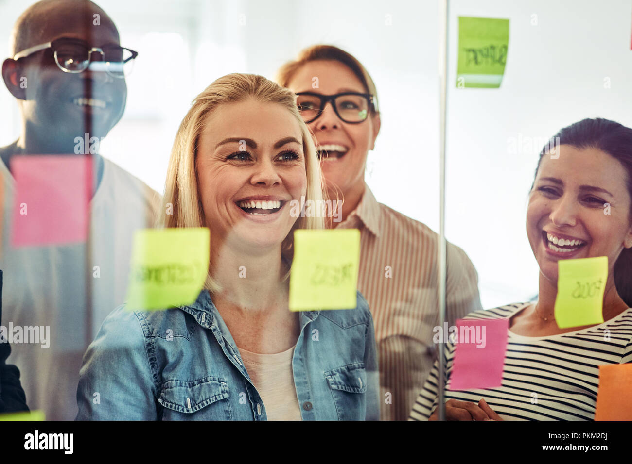 Ridendo gruppo di diversi imprenditori il brainstorming con foglietti adesivi su una parete di vetro mentre in piedi insieme in un ufficio moderno Foto Stock