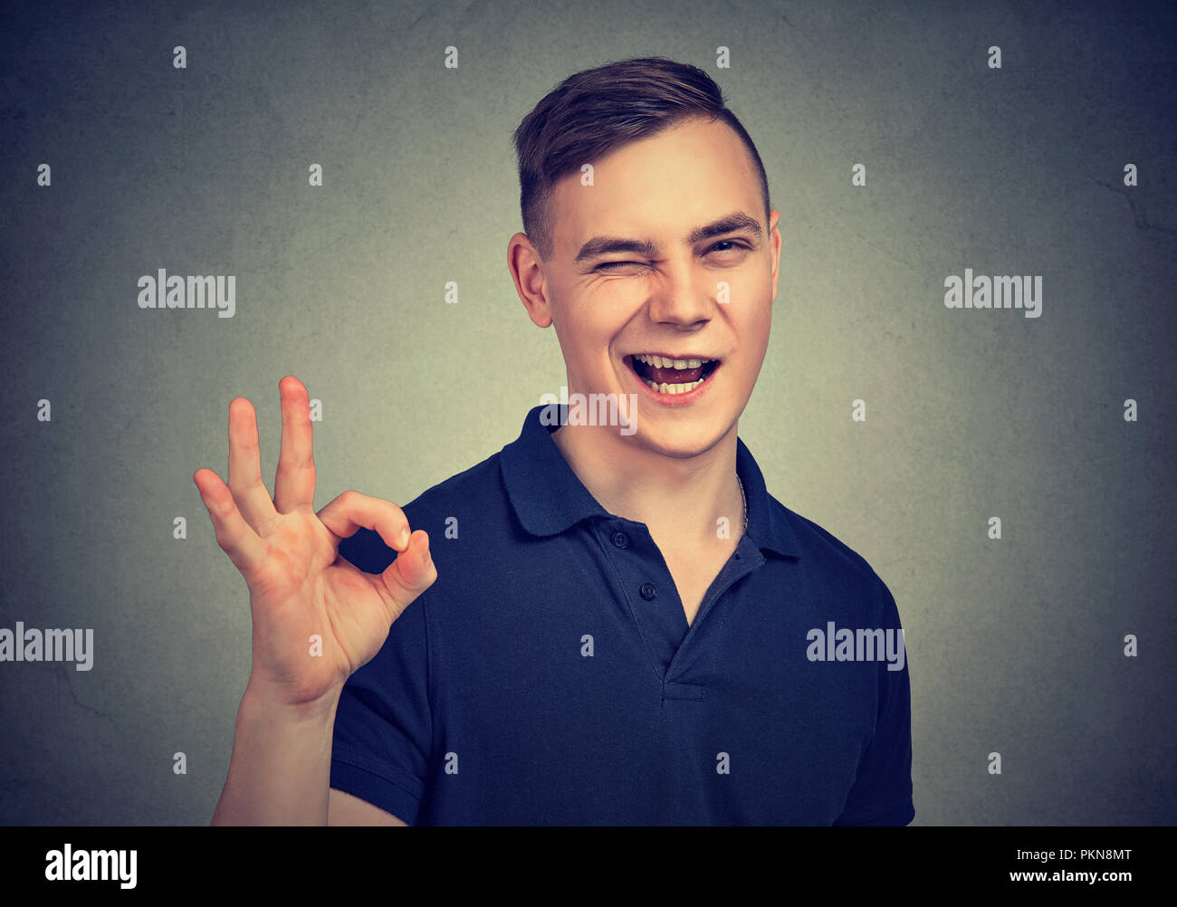 Bel uomo informale con una strizzatina d'occhio alla telecamera e mostra OK gesto su sfondo grigio Foto Stock