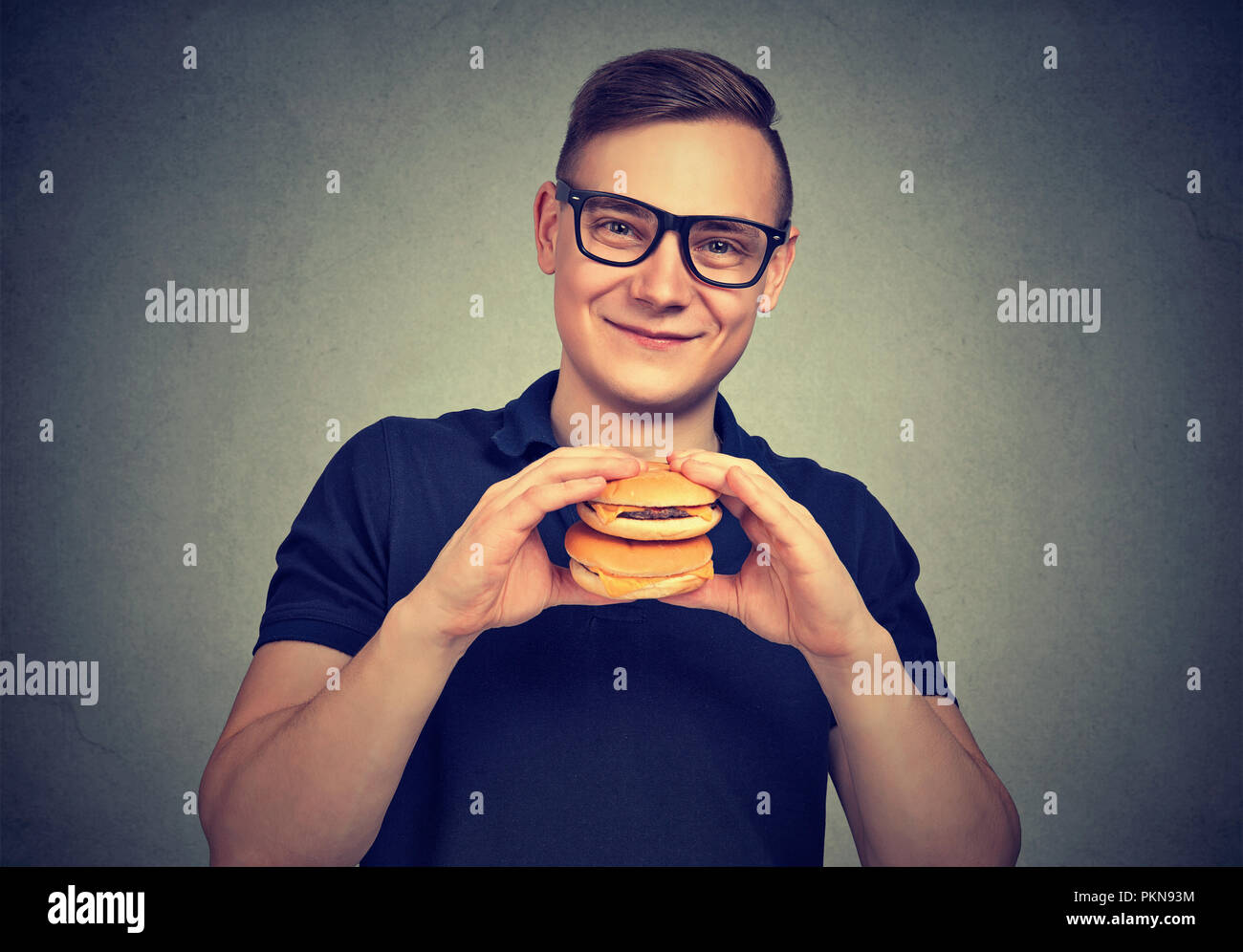 Giovane uomo sorridente in bicchieri holding alcuni hamburger e sorridente in telecamera godendo il fast food Foto Stock