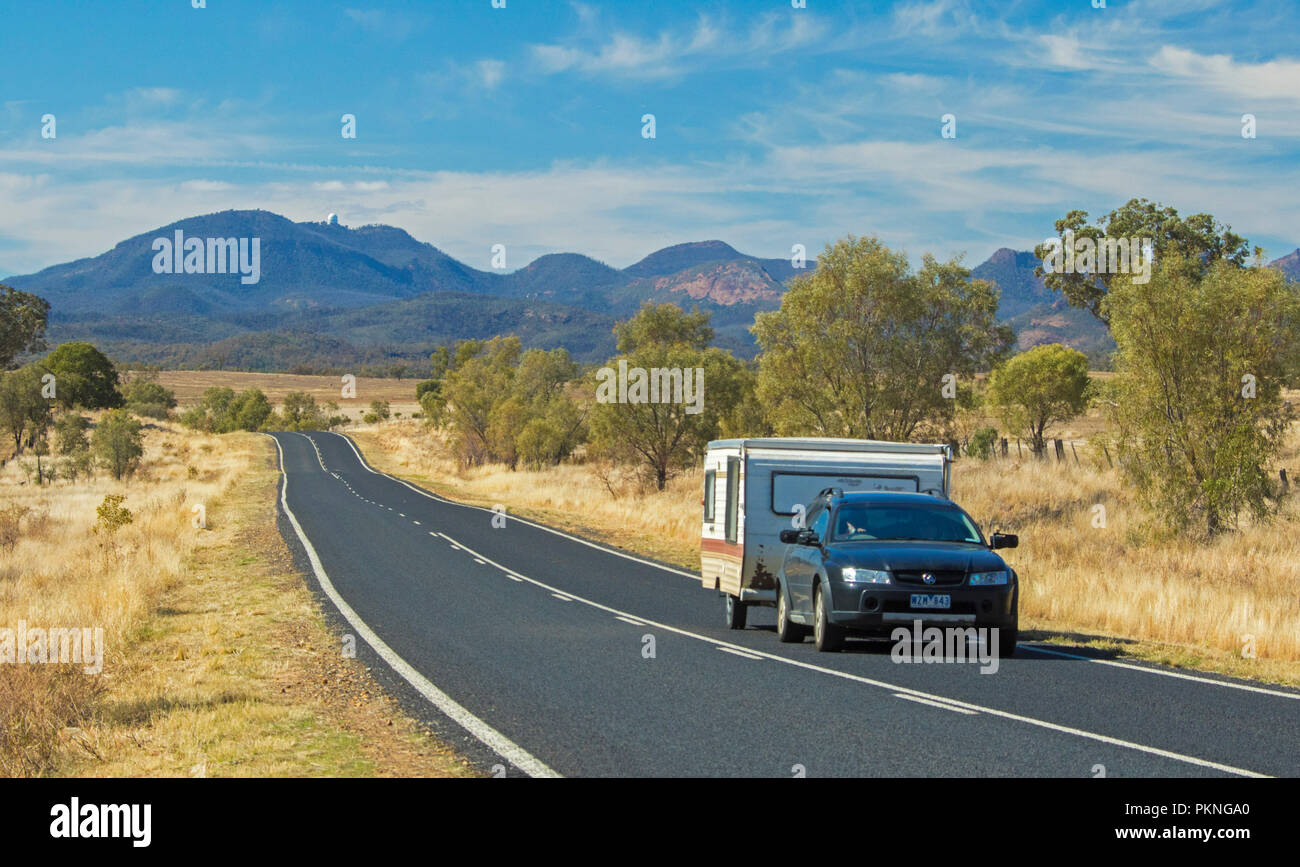 Auto con roulotte su strada che conduce attraverso il paesaggio di golden erbe & boschi a picchi di Warrumbungle National Park salga nel cielo blu in NSW Foto Stock