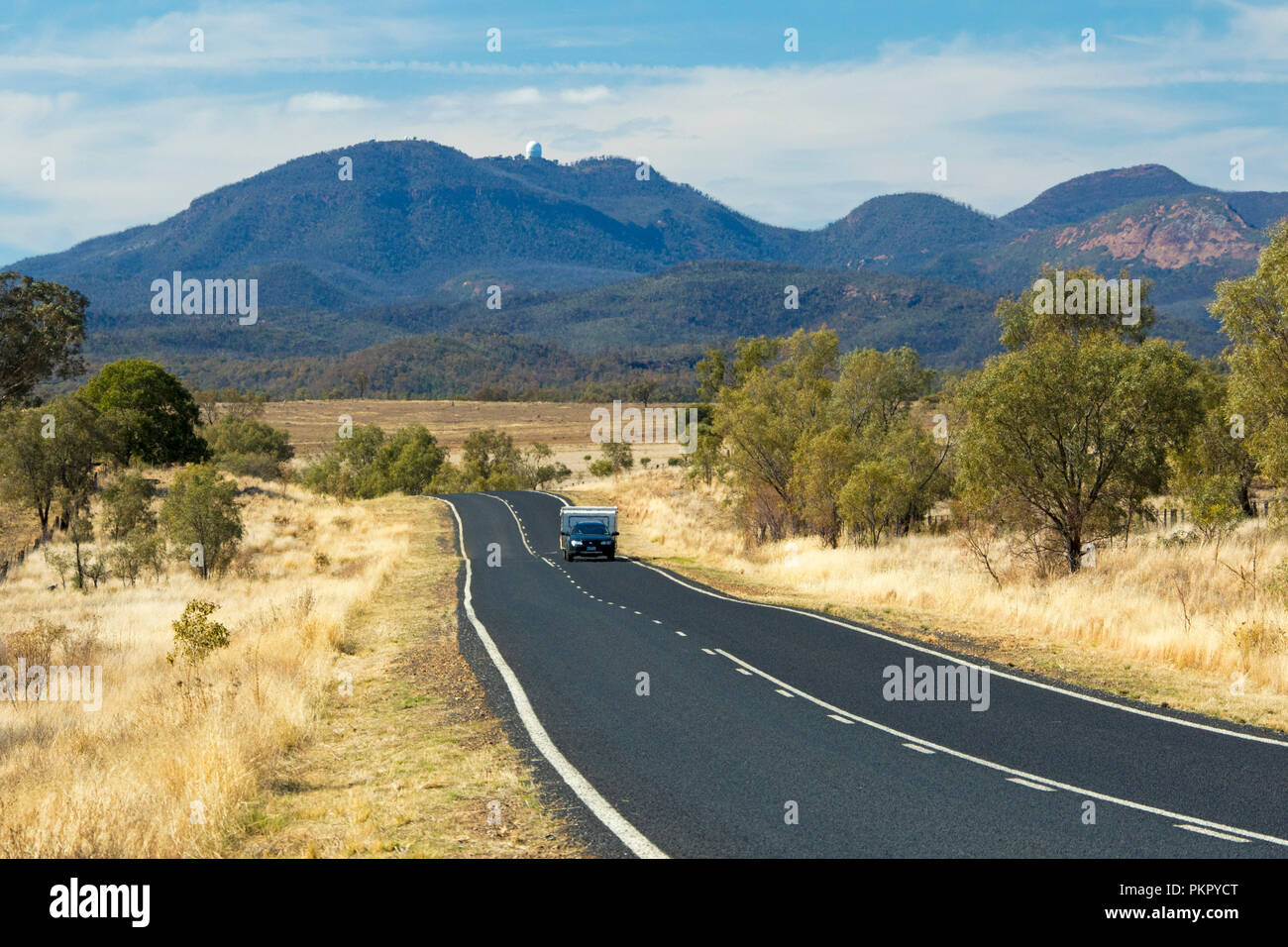 Auto con roulotte su strada che conduce attraverso il paesaggio di golden erbe & boschi a picchi di Warrumbungle National Park salga nel cielo blu in NSW Foto Stock