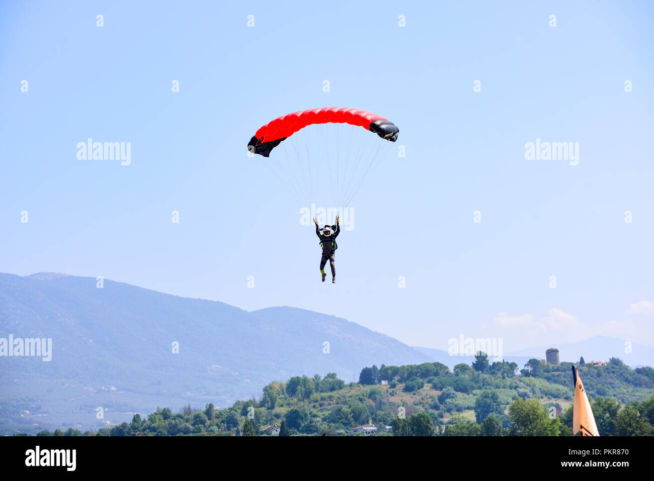 Paracadutista con paracadute rosso ottenere pronto per l'atterraggio. Su uno sfondo di cielo blu, montagne e colline verdi Foto Stock