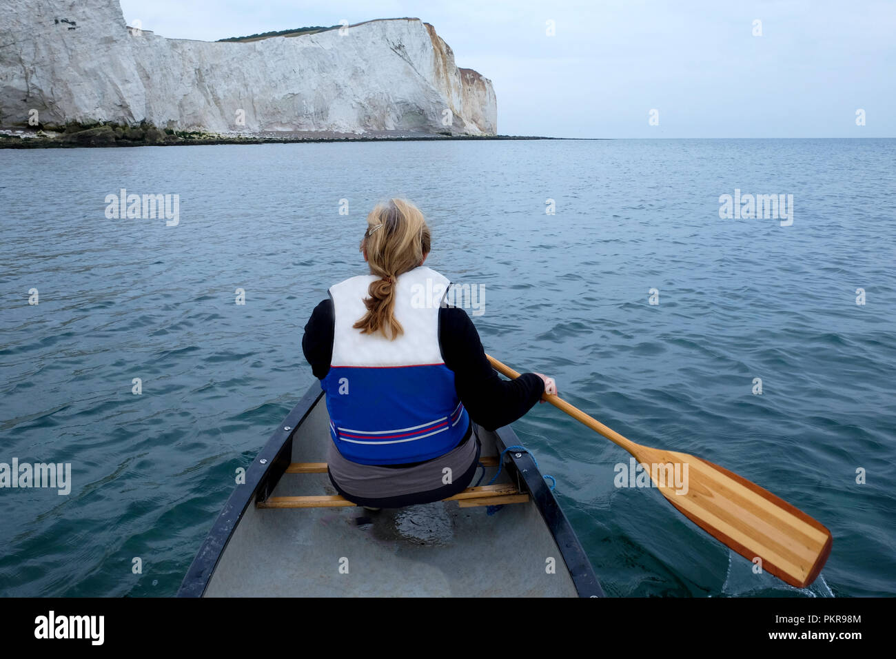 La vista posteriore di una sessanta anni di donna seduta nella parte anteriore di un kayak paddling con una paletta di legno indossa un giubbotto di salvataggio e ha capelli biondi Foto Stock