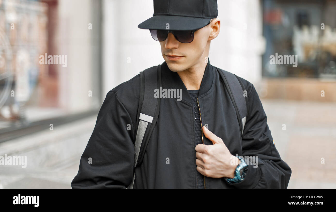 Giovane uomo di moda occhiali da sole in un cappuccio nero e giacca con un sacchetto in città Foto Stock