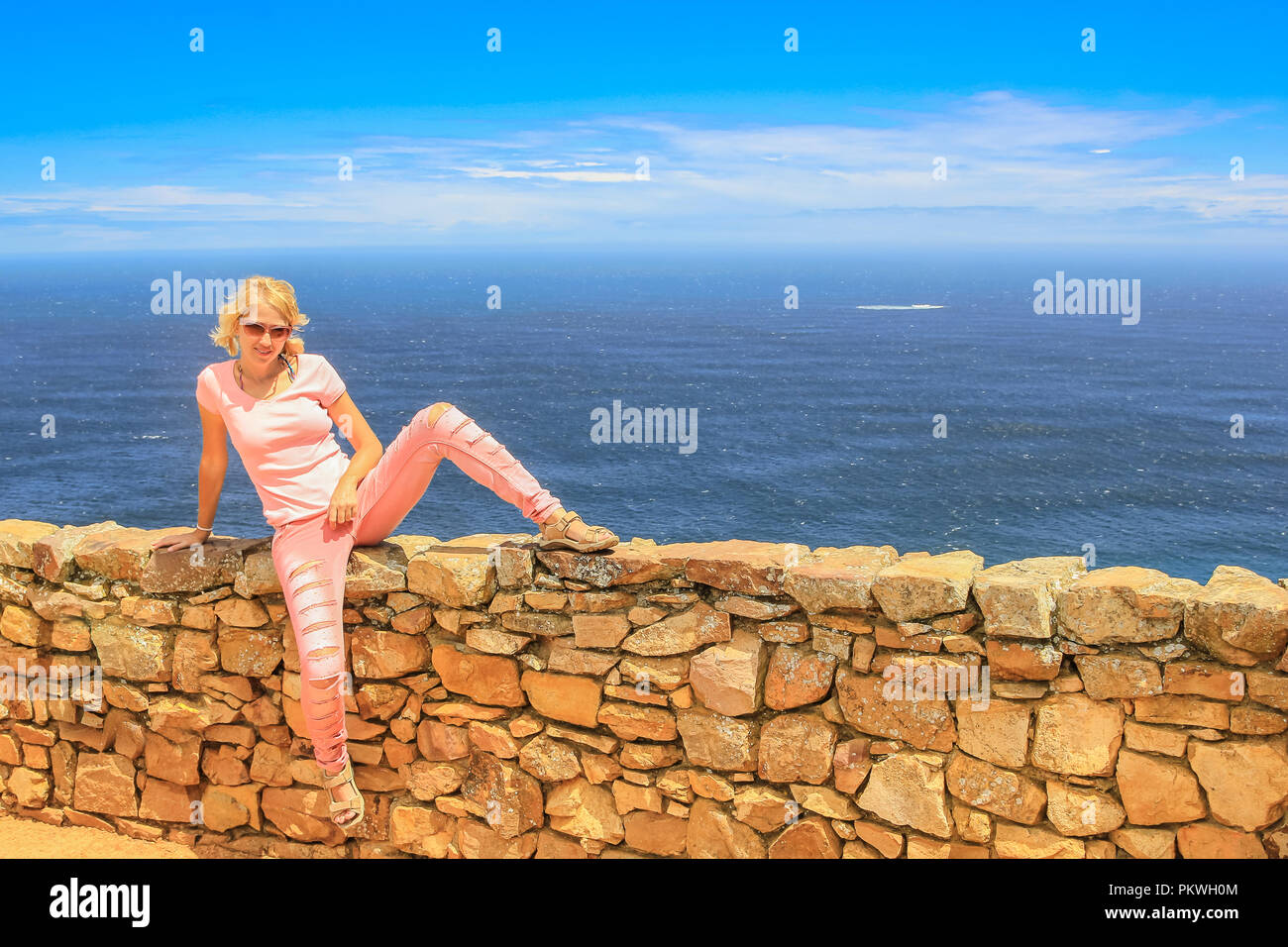Stile di vita turista donna seduta su un basso muro lungo il sentiero che conduce a Cape Point Lighthouse in Capo di Buona Speranza, Cape Peninsula, Western Cape. Turista femminile con oceano Atlantico sullo sfondo. Foto Stock