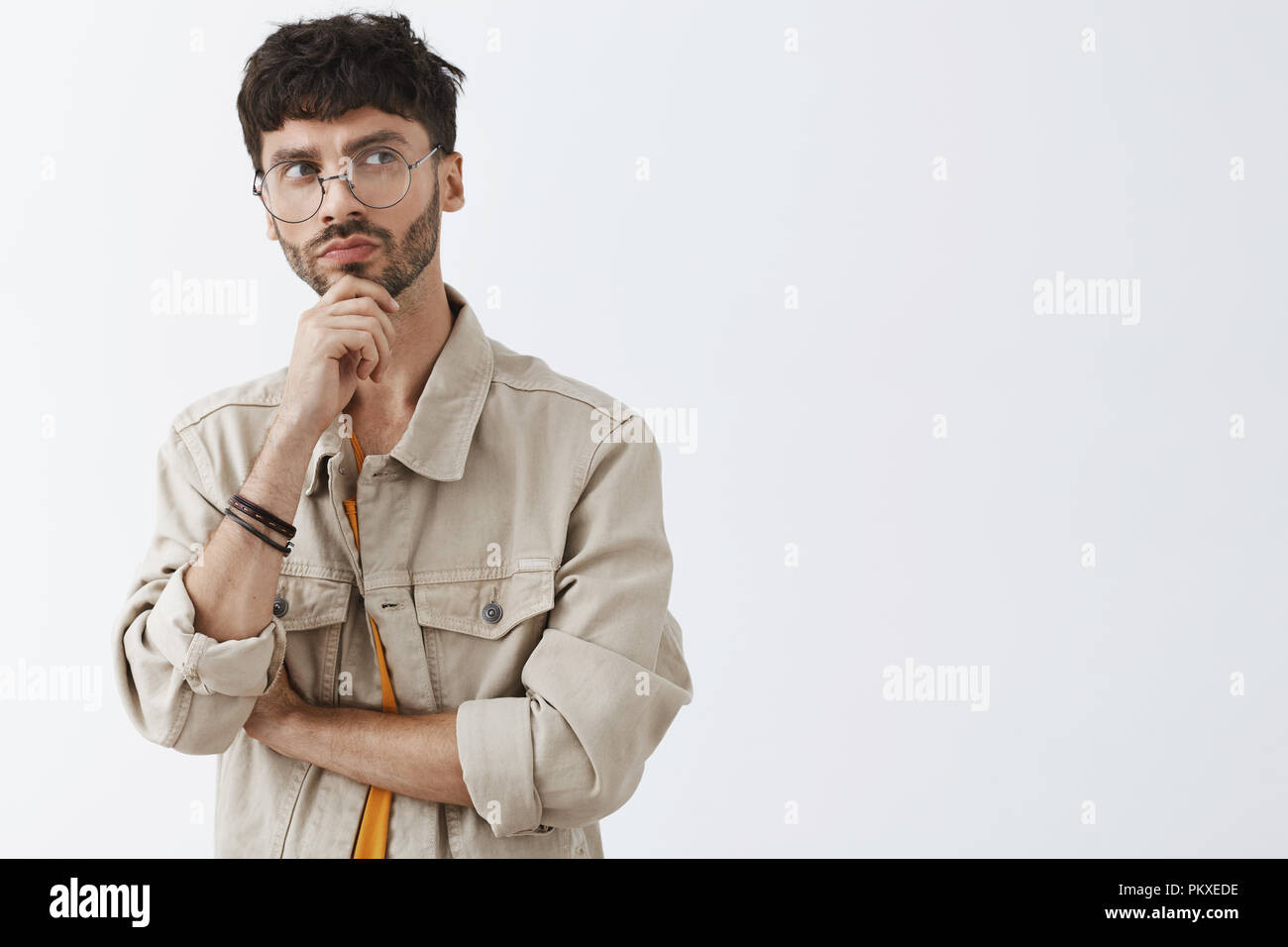 Vita-up shot di europeo creativo dai capelli scuri con uomo in barba alla moda di occhiali e maglietta beige tenendo la mano sul mento e guardando a destra con premurosa e espressione strategica oltre il muro grigio Foto Stock